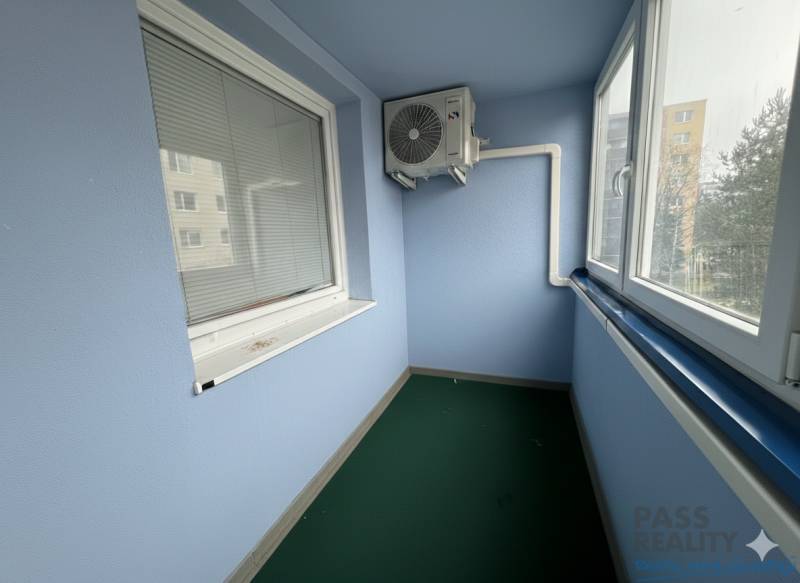 Zasklená lodžie s klimatizační jednotkou v 2-pokojovém bytě, vybavená žaluziemi a zeleným kobercem.