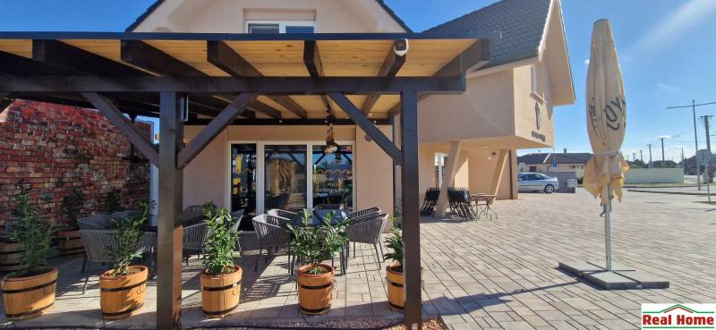 Gastro prostory v Nové Dědince s pergolou, květináči a venkovním posezením na dlažbě.