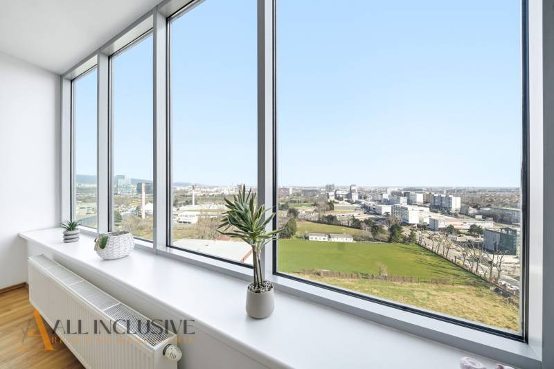 Výhled z 2pokojového bytu s velkými okny, pokojové rostliny, podlaha s dřevěným dekorem.