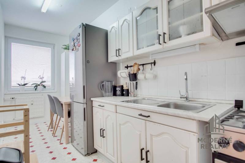 Kuchyně v 3-pokojovém bytě s bílými skříňkami, jídelním stolem a velkou lednicí.