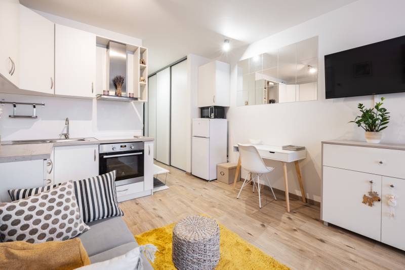 Obývací pokoj jednopokojového bytu s kuchyňskou linkou, bílým nábytkem a podlahou s dřevěným dekorem.