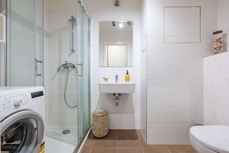 Koupelna v 1-pokojovém bytě s pračkou, sprchovým koutem a bílými obklady.