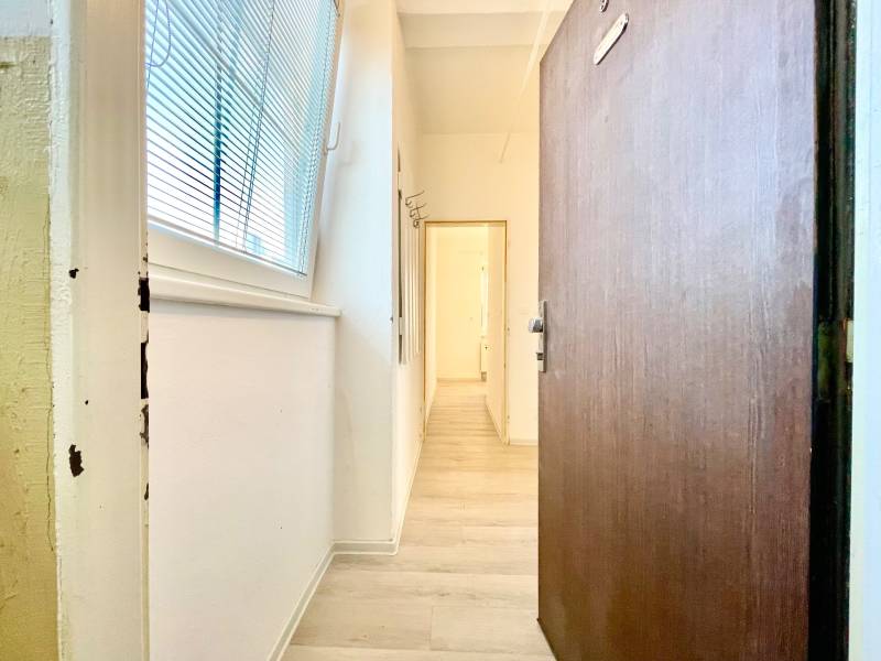 Chodba s podlahou s dřevěným dekorem, bílé stěny, velké okno se žaluziemi.