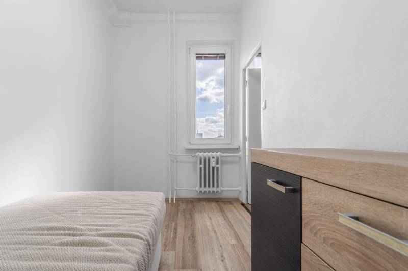 Jednoduchá izba s posteľou, komodou a podlahou s dreveným dekorom v 1-izbovom byte.