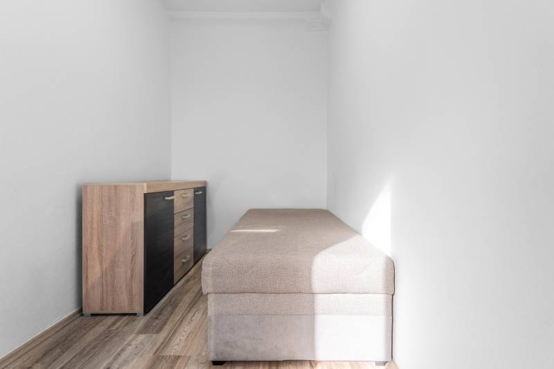 Úzka miestnosť s posteľou a komodou, podlaha s dreveným dekorom, 1-izbový byt.