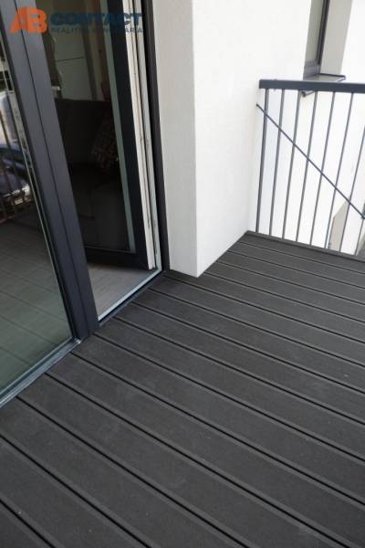 Balkon s dřevěnou podlahou na ulici Bodona, součást garsonky s velkým oknem.