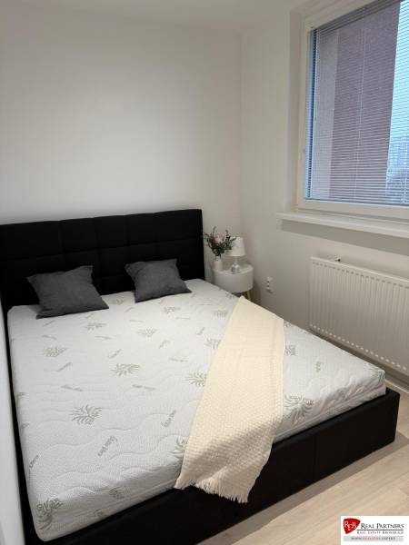 Ložnice v dvoupokojovém bytě s černým čelem postele, bílou přikrývkou a oknem s roletami.