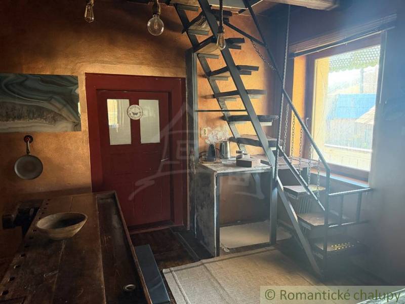 Interiér rodinného domu s kovovým schodištěm a dřevěným stolem, světlé stěny.