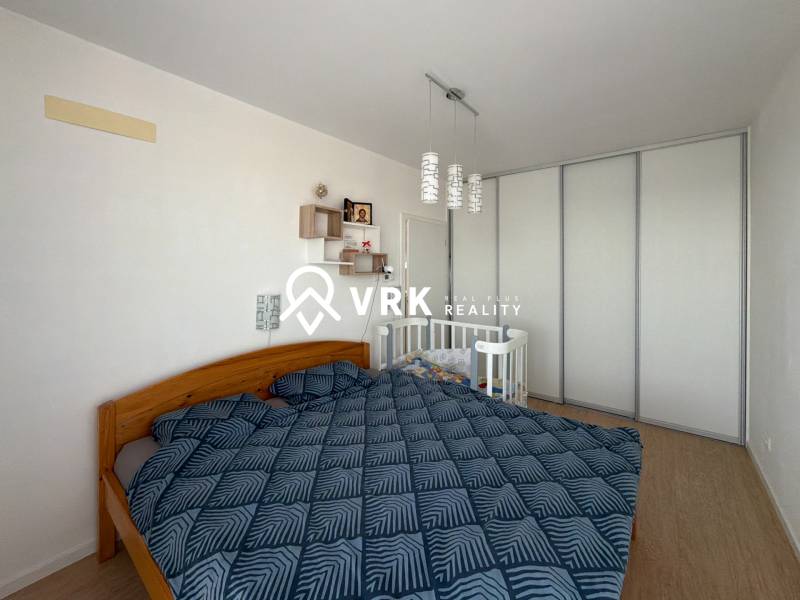 Ložnice v třípokojovém bytě s manželskou postelí, dětskou postýlkou a podlahou s dřevěným dekorem.