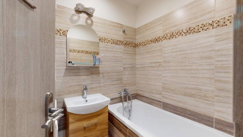 Koupelna v 2-pokojovém bytě se světlými obklady a vanou, skříňky pod umyvadlem.