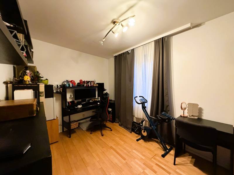 Pokoj s podlahou s dřevěným dekorem, psací stůl, počítač, stacionární kolo, police.