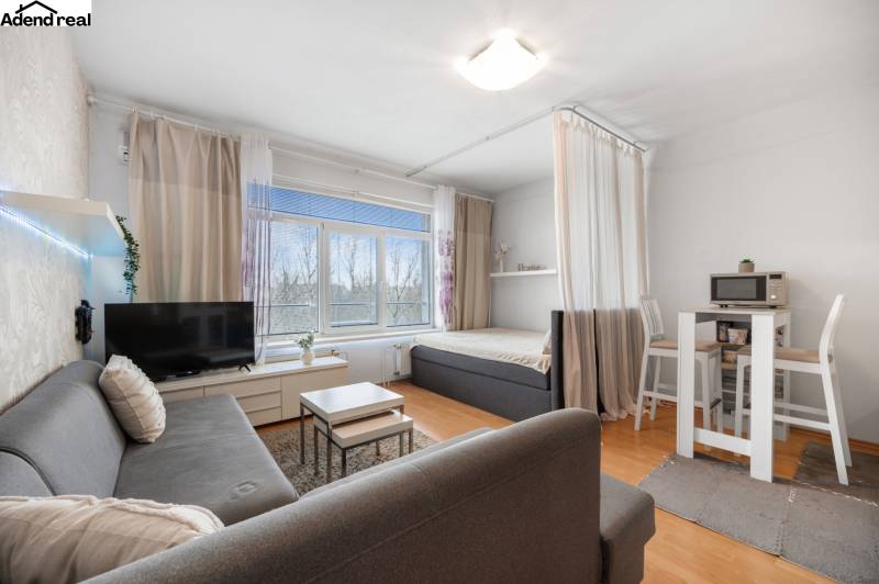 Obývací část 1-pokojového bytu s pohovkou, TV, jídelním stolem a postelí.