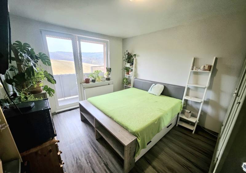 Světlá ložnice s postelí, rostlinami a podlahou s dřevěným dekorem ve 3-pokojovém bytě.