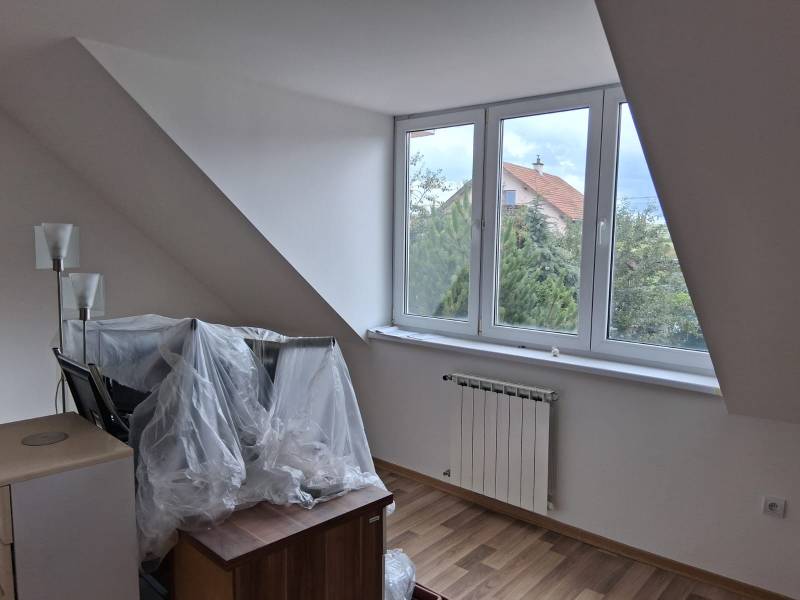 Podkrovní pokoj s velkým oknem, radiátorem a podlahou s dřevěným dekorem.