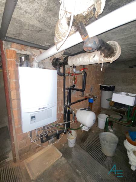 Plynový kotel ve sklepě rodinného domu s potrubím a zásobníkem vody na stěně.