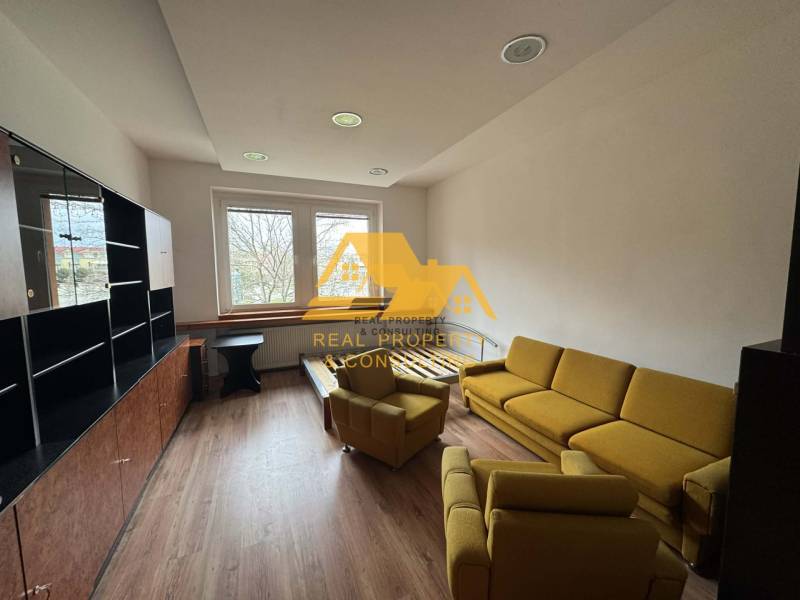 Obývací pokoj v 1+kk bytě s podlahou s dřevěným dekorem, žlutou sedačkou a velkým oknem.