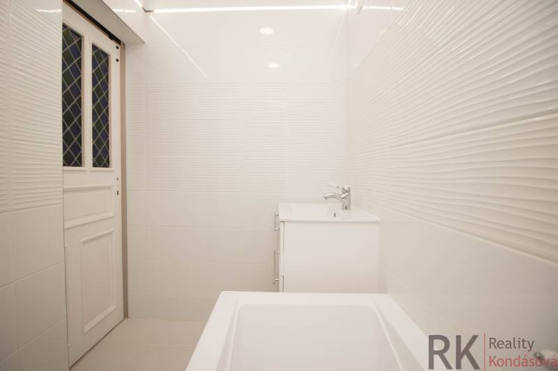 Koupelna v 2-pokojovém bytě s bílými obklady, umyvadlem a vanou.