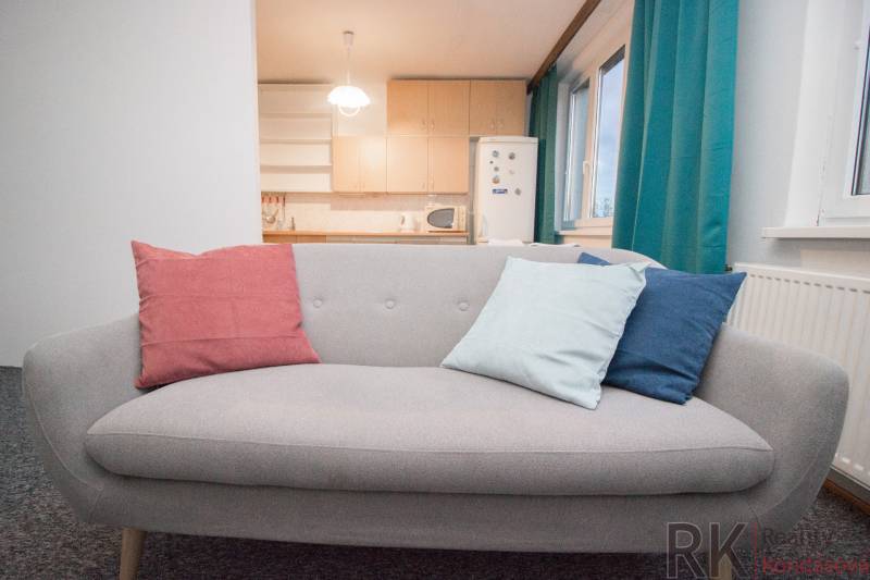 Šedá pohovka s barevnými polštáři v 4pokojovém bytě u okna a kuchyně.