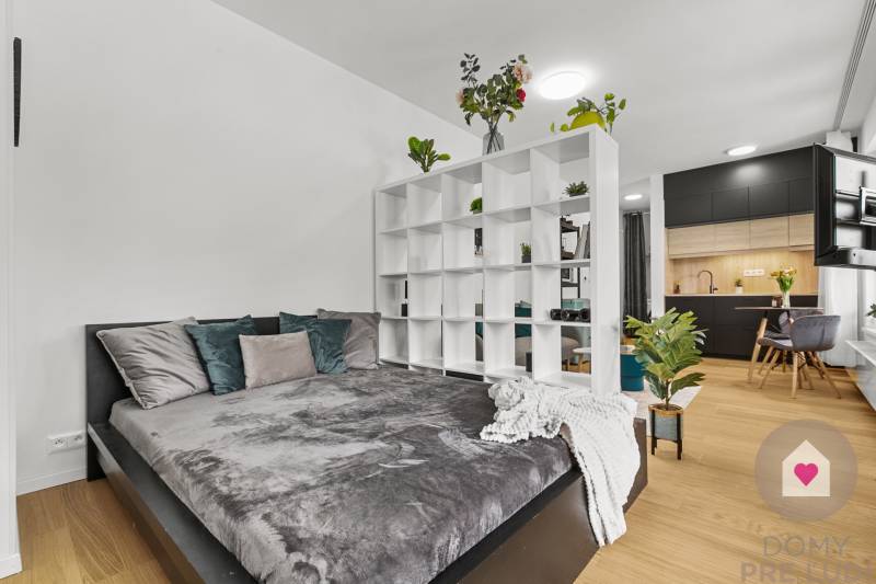 1-pokojový byt s postelí, regálem, podlahou s dřevěným dekorem a černou kuchyní.