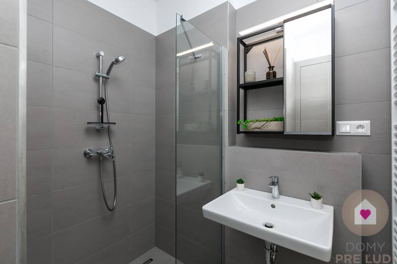 Šedá koupelna se sprchou, umyvadlem a velkým zrcadlem v 1-pokojovém bytě.