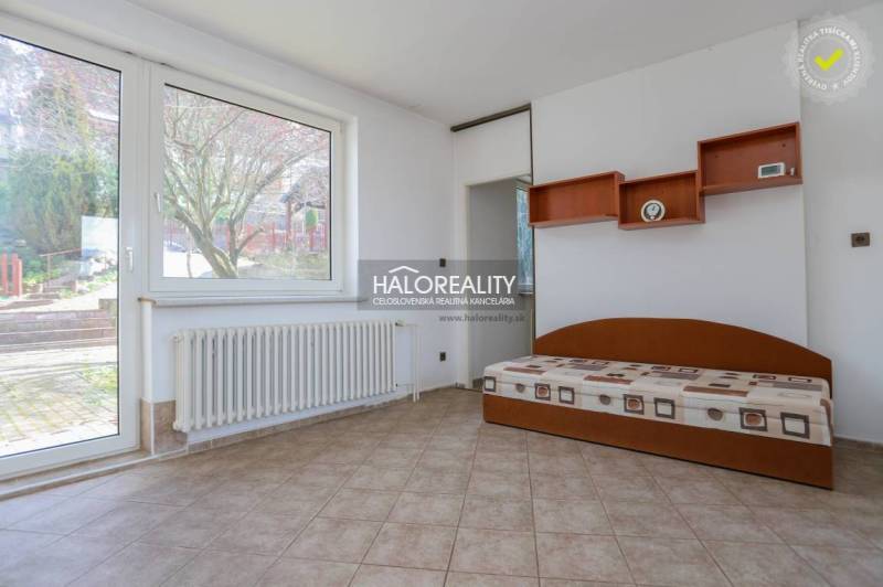 Interiér rodinného domu s postelí, poličkami, radiátorem a velkým oknem.