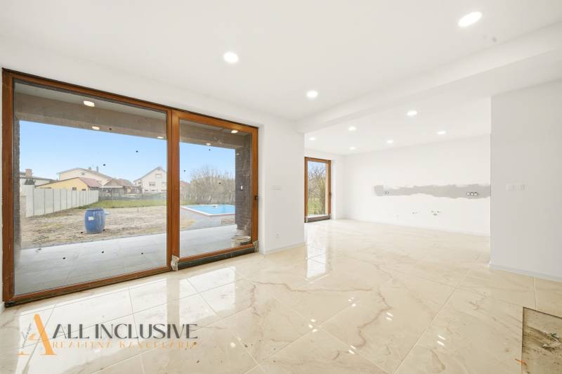 Prostorný interiér rodinného domu s velkými okny a výhledem na zahradu s bazénem.