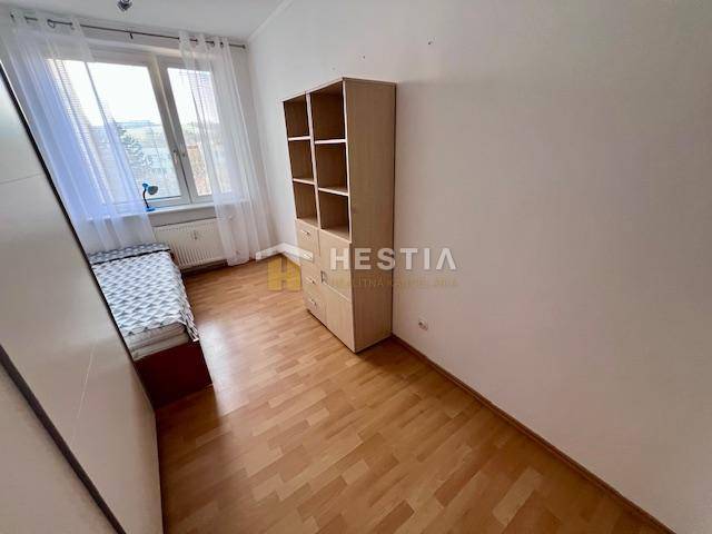 2-pokojový byt, postel u okna, regál, podlaha s dřevěným dekorem, bílá stěna.