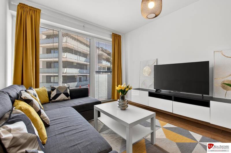 Dvoupokojový byt s velkými okny, pohovkou, televizí, žlutými závěsy a podlahou s dřevěným dekorem.