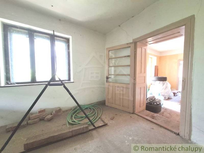 Pokoj v chatě se světlem z okna, otevřenými dveřmi a stavebním nepořádkem.