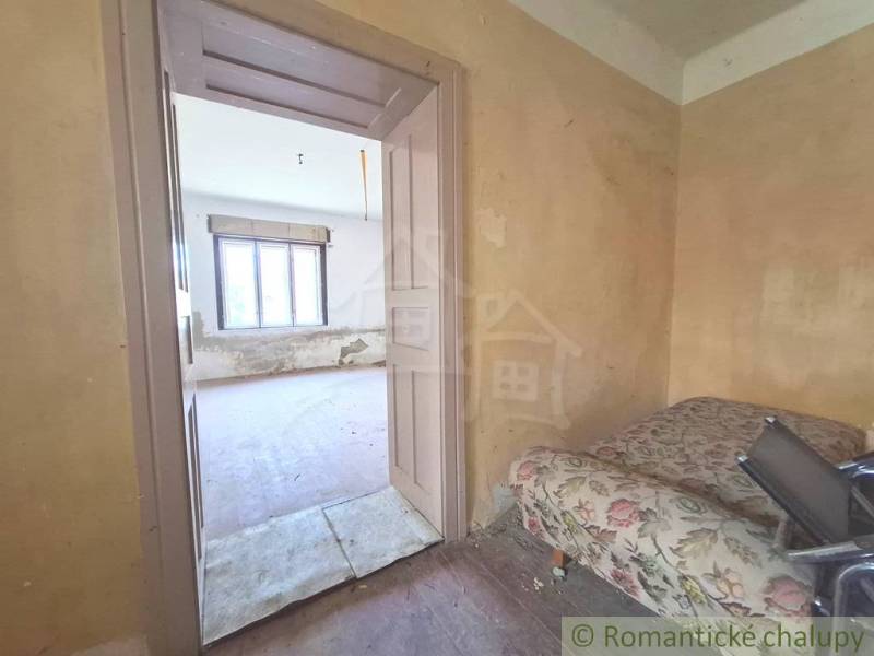 Pokoj na chatě se starým nábytkem a oknem, čeká na renovaci.