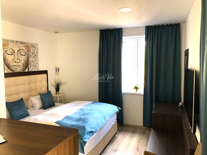 Garsonka se světlým nábytkem, postelí a modrými závěsy, podlaha s dřevěným dekorem.