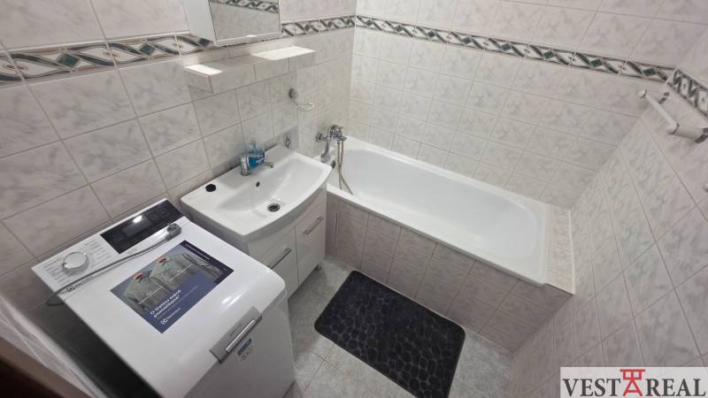 Koupelna s vanou a pračkou v 1-pokojovém bytě, světlé obklady a úložný prostor.