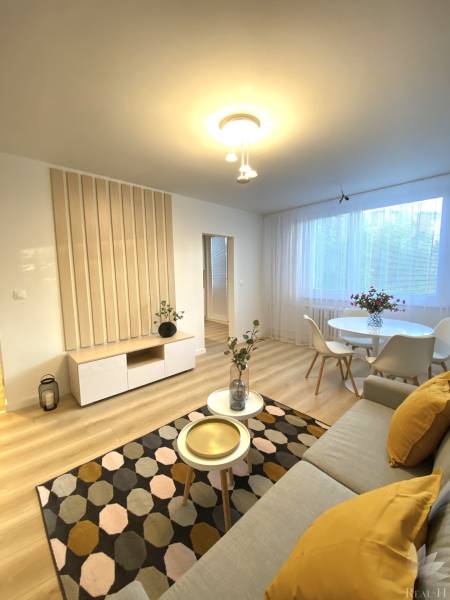 Obývací pokoj dvoupokojového bytu s podlahou s dřevěným dekorem, světlým nábytkem a dekoracemi.