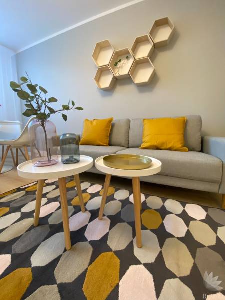 Obývací pokoj v 2-pokojovém bytě s geometrickými poličkami, šedou sedačkou a barevným kobercem.