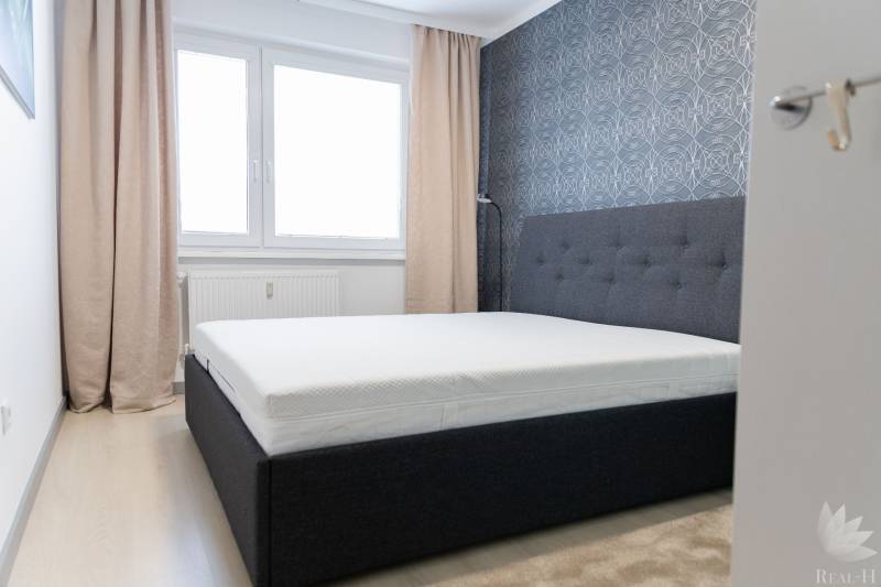 Ložnice ve 4pokojovém bytě s postelí, tapetou, okny a závěsy, podlaha s dřevěným dekorem.