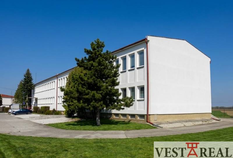 Budova v Šoporni obklopená zelení a stromy, s jasnou modrou oblohou.