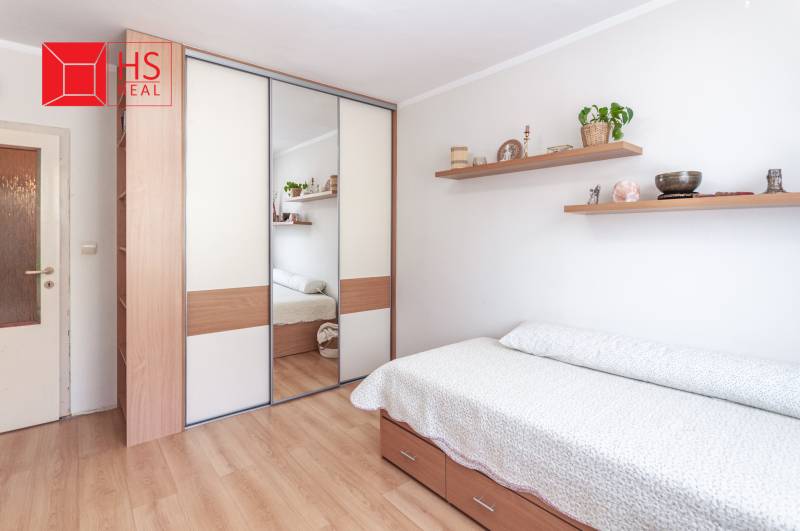 Ložnice v třípokojovém bytě s postelí a zrcadlovou skříní, podlaha s dřevěným dekorem.