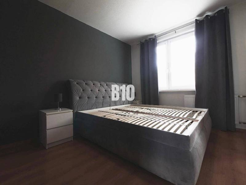 Ložnice s tmavě šedou postelí, nočním stolkem a podlahou s dřevěným dekorem ve 3pokojovém bytě.