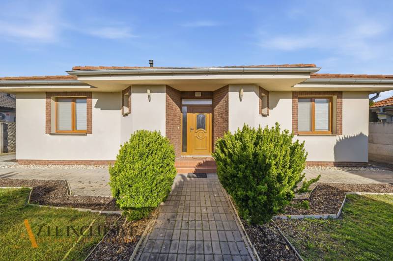 Rodinný dům v Kútnikách s keřovou výsadbou, bílou fasádou a hnědými doplňky.