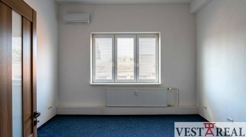 Místnost s klimatizací a oknem v Kancelářích, modrý koberec, bílé stěny.