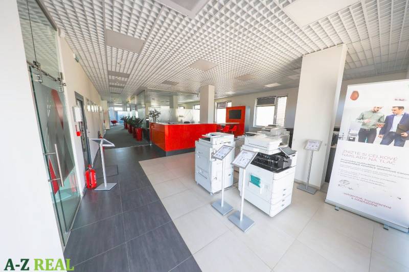 Interiér kanceláře s tiskárnami, červeným recepčním pultem a vysokým stropem.