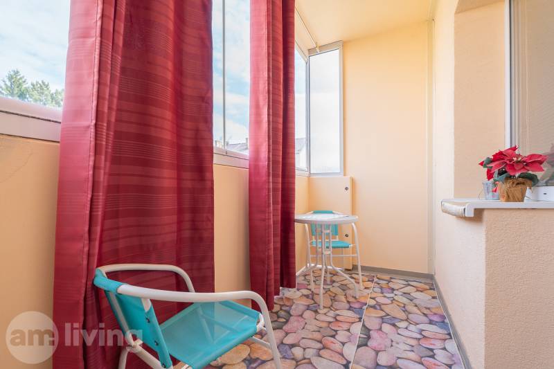 Balkon v 1-pokojovém bytě s červenými závěsy, stolkem a rostlinou na parapetu.