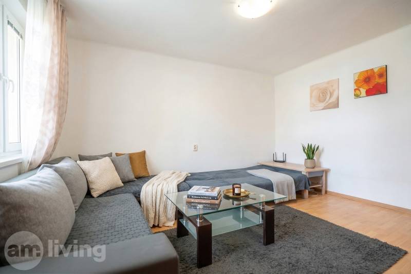 Světlý 1-pokojový byt s rohovou sedačkou, skleněným stolkem a podlahou s dřevěným dekorem.