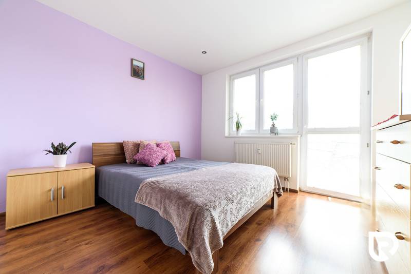 Ložnice v dvoupokojovém bytě s postelí, skříní a podlahou s dřevěným dekorem.