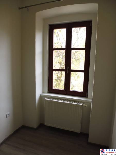 Pokoj s velkým oknem, radiátorem a podlahou s dřevěným dekorem ve 3-pokojovém bytě.