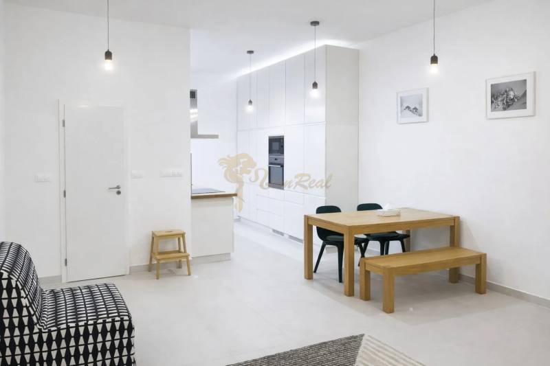 Interiér 2-pokojového bytu s bílými stěnami, dřevěným stolem a černou sedačkou.