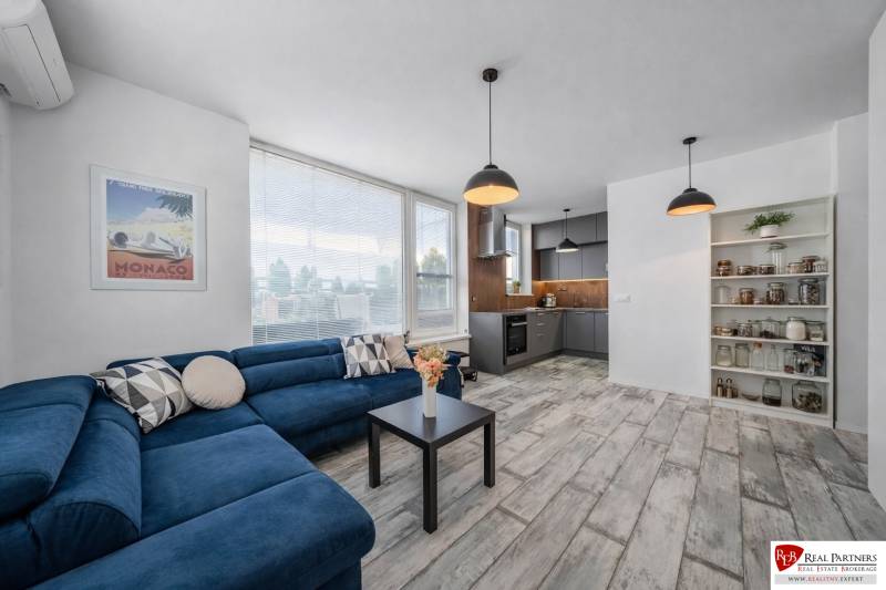 Obývací pokoj s podlahou s dřevěným dekorem, modrou sedačkou a kuchyňským koutem v 2-pokojovém bytě.