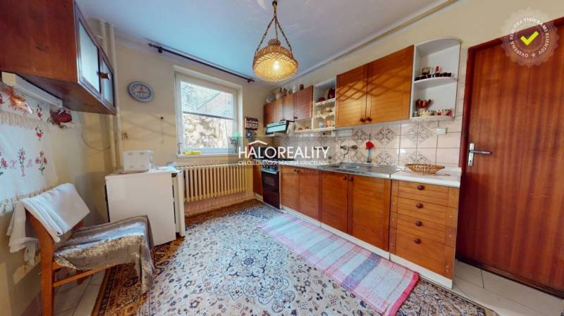 Kuchyně v rodinném domě s dřevěnými skříňkami, kobercem a závěsným svítidlem.