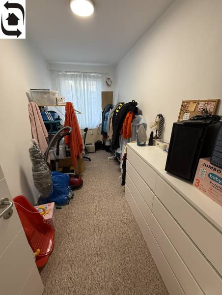 Úzká místnost ve 4-pokojovém bytě s komodou, regály a oknem bez záclon.