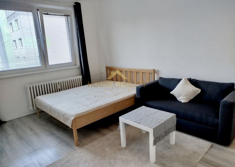 Pokoj s postelí, černou pohovkou a stolkem, podlaha s dřevěným dekorem.